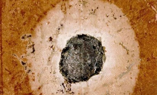 这块嵌在石灰岩中的、已经化石化的陨石在欣讷山十分典型。正中间的一团黑色物质便是陨石本身，陨石周围的灰色区域则是被氧化成红色的石灰石在受到陨石撞击时发生了还原反应