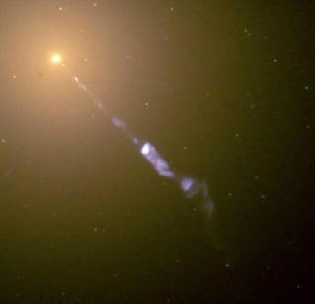 M87星系的中心闪耀着一个巨大的宇宙探照灯：从黑洞向外喷射出的次原子粒子流的速度接近光速。 在这张哈伯太空望远镜所拍摄的影像中，蓝色的喷流与M87星系中恒星和星